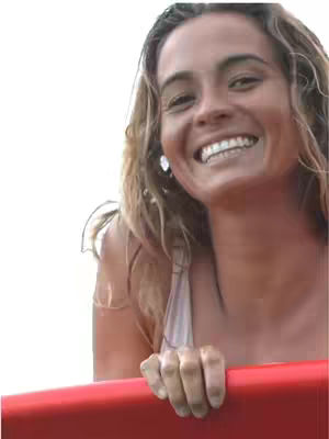 A woman with long wavy light brown hair smiles brightly, holding onto a red bar. The background is a blurred white.