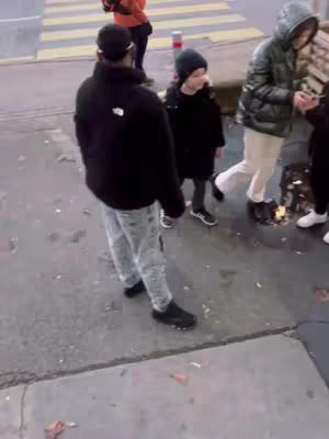 The image shows a street scene, with a group of people walking. The foreground features a person wearing a black jacket and grey pants, their back facing the camera. A child in black clothing and a beanie stands nearby, and another person in a green jacket walks past, next to a concrete curb. The background includes a crosswalk and some other individuals.