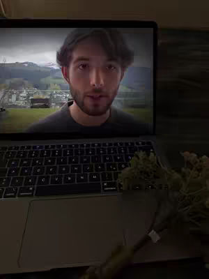 A man is featured on the screen of a laptop, set against the backdrop of a scenic landscape visible through the laptop's display. Beside the laptop, a bouquet of dried flowers add a textural contrast to the tech-focused setting.
