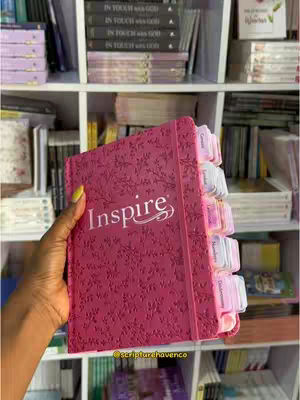 A person's hand holding a pink book titled "Inspire" in front of a shelf filled with books. The book has a floral embossed design and color-coded tabs. The background is a bookshelf with various books and items.