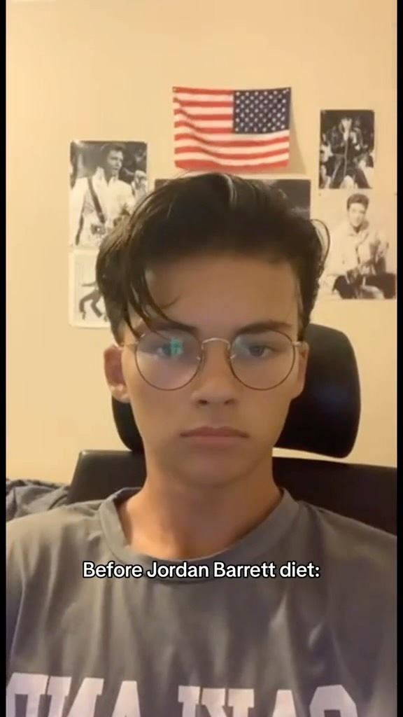 A close-up shot of a young man with dark hair, round glasses, and a neutral expression. He is wearing a gray t-shirt, and the background shows posters and an American flag.