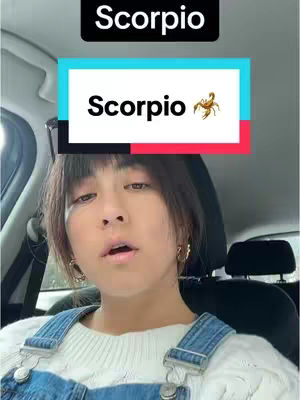 A woman is shown inside a car, looking at the camera. Above her head is a rectangle that is half black and half red with the word "Scorpio" in white font. Below this is a rectangle with a cyan and red border, the word "Scorpio" in white font, and a graphic of a scorpion. She is wearing denim overalls and a white sweater. The background is the interior of a car.