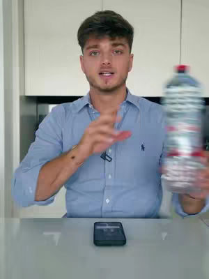 A man stands in front of a kitchen setting, holding a blurred water bottle in his right hand. He gestures with his left hand towards a phone lying on the table in front of him.