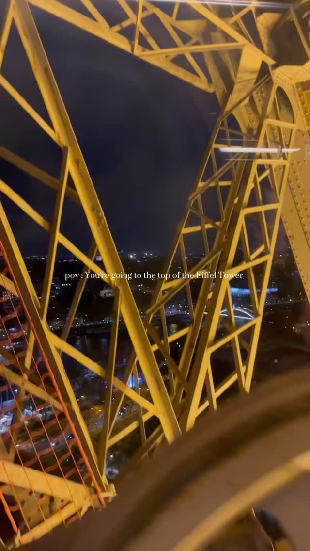 An interior shot from inside the Eiffel Tower with a view of the iron structure and night sky. The yellow iron framework of the tower's structure dominates the foreground, with visible joins and support beams creating a complex geometric pattern. The dark night sky serves as a backdrop, with city lights faintly visible in the distance. Text overlay indicates the video is about going to the top of the Eiffel Tower.