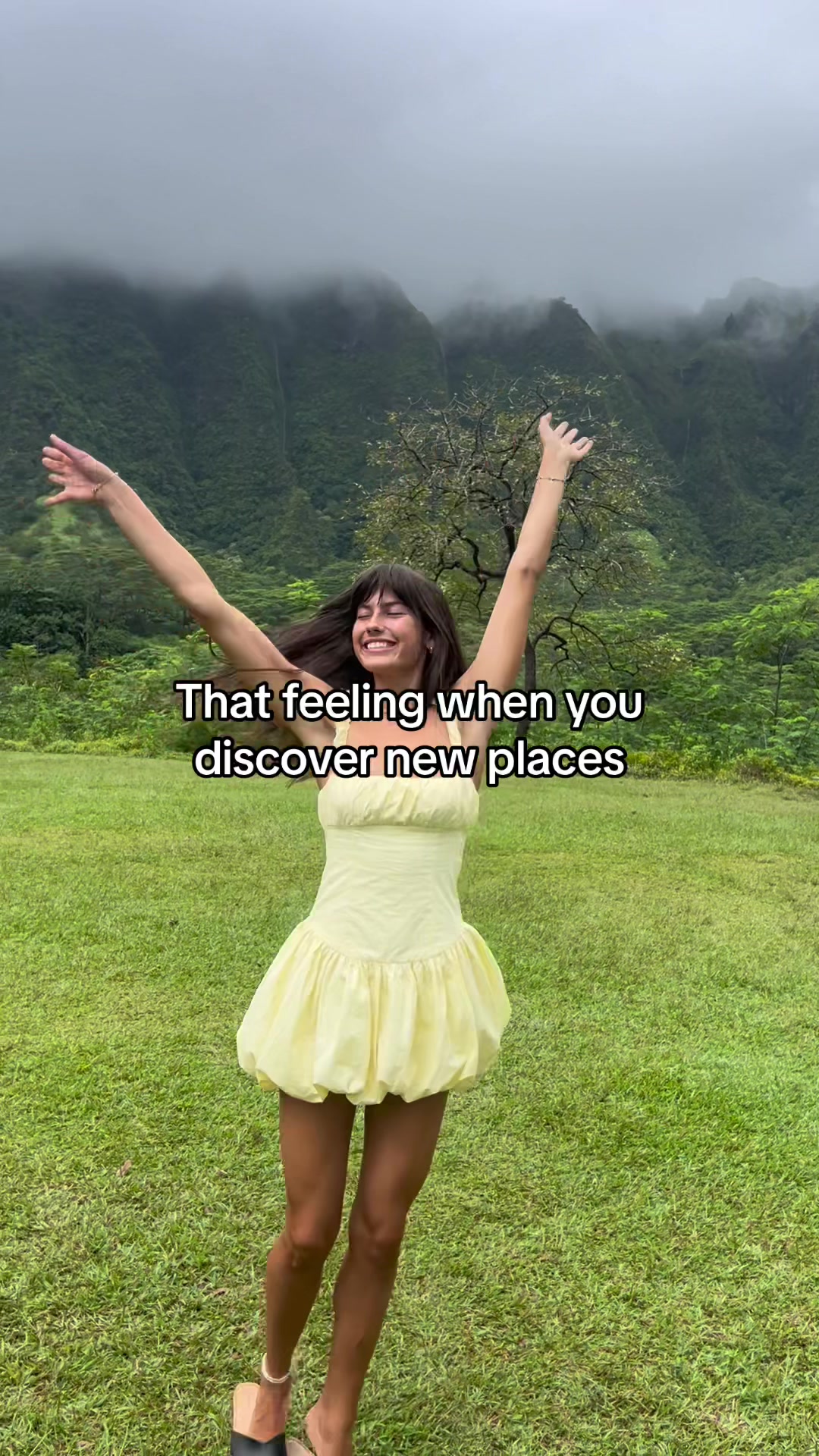 A woman with long dark hair and bangs stands in a grassy field with her arms outstretched, the background shows a lush mountainous landscape under an overcast sky. The woman wears a yellow dress. Text overlayed reads 'That feeling when you discover new places.'