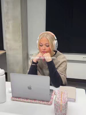 A young woman with blonde hair wearing a beige sweater and headphones sits at a desk with a laptop, her hands clasped under her chin as she stares at the screen. The laptop is open, and accessories like a notebook and a floral patterned pouch are visible. Soft, natural light suggests a daytime setting. Text overlay reads 'just because it burns'.