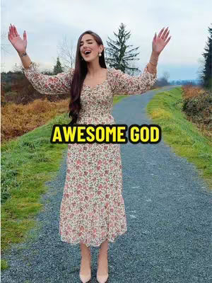 A woman stands on a gravel path with arms raised to the sky, the words "AWESOME GOD" are overlaid in text. The scene is set outdoors with a blurred background of trees and a cloudy sky.
