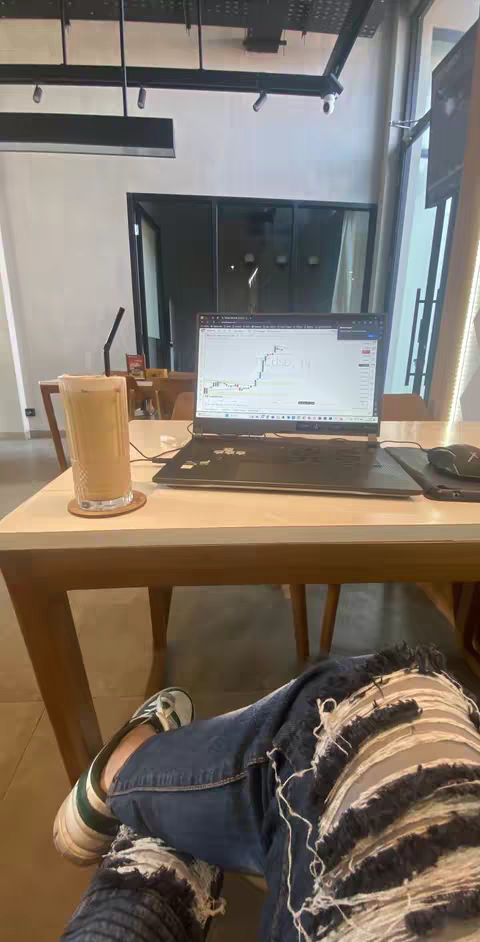 The image shows a person's legs and feet resting on a table in a café setting. The person is wearing distressed denim jeans and green and white sneakers. On the table in front of them sits a laptop displaying a stock chart, a glass of iced coffee, and a mouse. The lighting appears to be natural, with soft shadows suggesting a relaxed atmosphere.