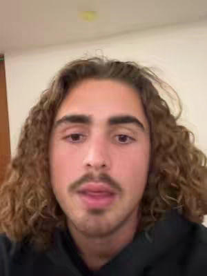 A young man with long curly brown hair and a mustache looks directly at the camera. He's wearing a black hoodie, and the background is a plain, light-colored wall. The lighting is neutral.