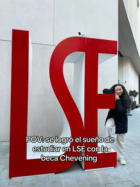 The image features a woman leaning against a large, red and white sculpture of the letters "LSE", which likely refers to the London School of Economics. The woman is smiling and appears to be happy. The text overlay reads "POV: Se logró el sueño de estudiar en LSE con la beca Chevening", translating to "POV: The dream of studying at LSE with the Chevening scholarship was achieved."