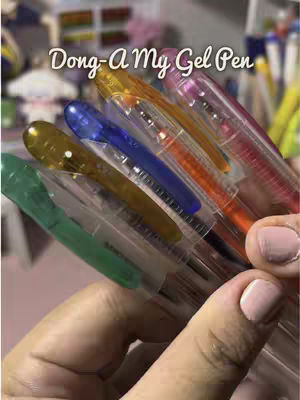 A hand holding a collection of colorful gel pens. The pens are tightly grouped together, with their transparent barrels revealing the ink colors within: green, yellow, blue, orange, and pink. The background is blurred, with hints of shelves and more stationery. The overall feel is bright and product-focused.