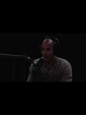 A man with a shaved head wearing a gray shirt and headphones is speaking into a microphone in a dimly lit room. The background is black. The man has a serious expression.
