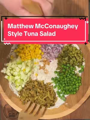 A overhead shot shows a wooden bowl filled with various ingredients for a tuna salad. The ingredients are arranged in sections around a central area, where the base of the salad appears to be. The sections include yellow corn, red onions, pickles, peas, and tuna, creating a colorful presentation. The hands are seen at top holding a knife.
