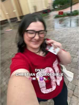 A selfie-style shot of a young woman smiling while holding a card, appearing to walk in the rain. She wears glasses, a red tee with 'DNTL' printed on it, and a white bag. The background show buildings, wet pavement, and foliage, implying an outdoor setting.