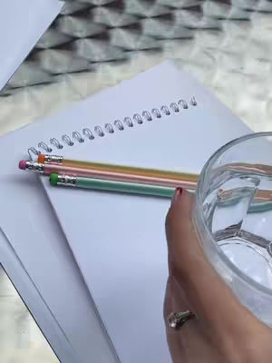 A hand holding a clear glass of water is in the foreground. Pencils and a notebook are positioned in the background. The composition is bright and well-lit.