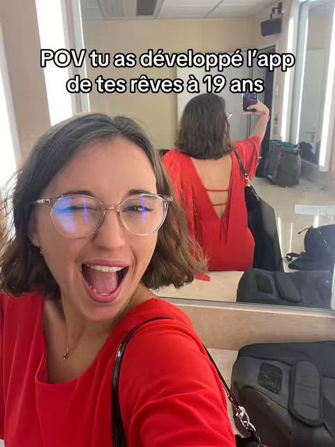 A selfie taken in a room with mirrors. The foreground features a woman smiling with her mouth wide open, wearing glasses and a red top. Behind her, in the mirror, a different view of the same woman is visible, taking a selfie wearing a red dress. Background elements include a dressing room setting with bags and other items.