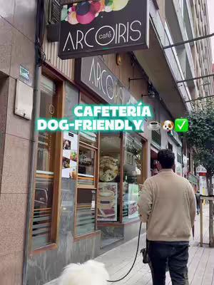 An eye-level exterior shot captures a man walking towards a cafe. The cafe's facade is visible, featuring a sign that reads 'ARCOIRIS cafe'. A vibrant text overlay declares the cafe 'CAFETERÍA DOG-FRIENDLY'. The man, dressed in a light-colored jacket and dark pants, is walking a dog on a leash. The dog, with white fur, is in front of the man. The background includes urban elements like buildings and a street.