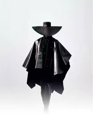 A full-body studio shot features a person in an all-black ensemble set against a stark white backdrop. The figure's most striking feature is an oversized, broad-brimmed hat that completely obscures the top of their head and casts a deep shadow over their face. They wear a structured, dark, cape-like garment that extends to their waist, partially concealing the torso. The lower body is less defined, possibly due to the clothing or shadow play, creating a sense of mystery. The high-key lighting emphasizes the textures of the clothing and the hat's shape, resulting in a bold, high-fashion or conceptual feel.
