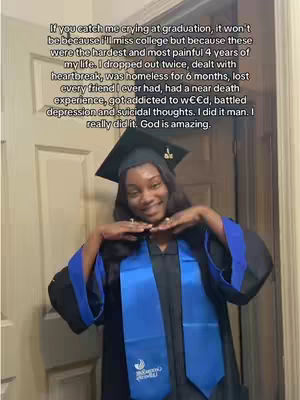 A young Black woman in a graduation gown and cap, smiling into the camera with both hands up near her face. She has long dark hair and is wearing a blue graduation sash. Behind her is a light-colored door and wall.