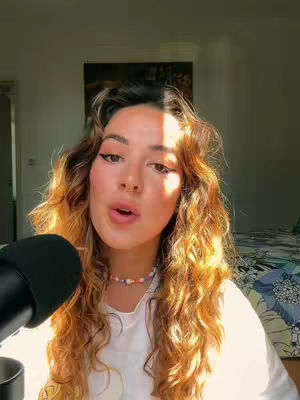 A young woman with long, wavy auburn hair and a white top is positioned in front of a microphone, with a bed and artwork visible in the background. She has eyeliner and a small necklace, and the lighting suggests a warm environment.