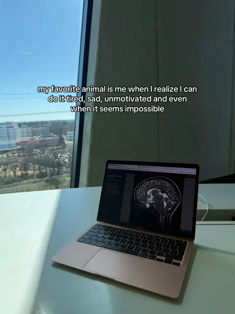 The image shows a laptop on a white desk near a large window. The laptop displays a brain scan, while the room has a bright and clean aesthetic. The caption states 'my favorite animal is me when I realize I can do it tired, sad, unmotivated and even when it seems impossible'.