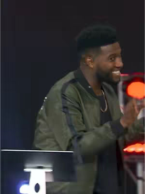 The cover image features a close-up of a Black man smiling while looking to the side. He is wearing a green bomber jacket over a black shirt and a gold necklace. The background is dark and partially obscured by shadows. A computer is visible in the foreground. The overall scene seems to be from a stage or studio setting illuminated by red lights.