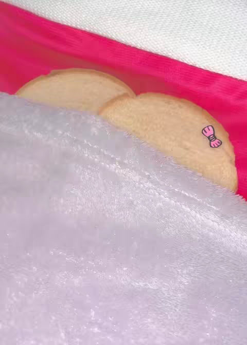 The image features two slices of bread, possibly cookies. They are light tan in color and partially covered by a fluffy, white blanket. A bright pink cloth provides a vibrant background. A small, pink bow sticker is on one slice.