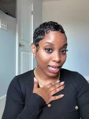 A woman with short, dark, curly hair poses in a selfie with a hand on her chest, expressing emotion. She wears a black top and a gold necklace. The background suggests an interior setting with a door and a light-colored wall.