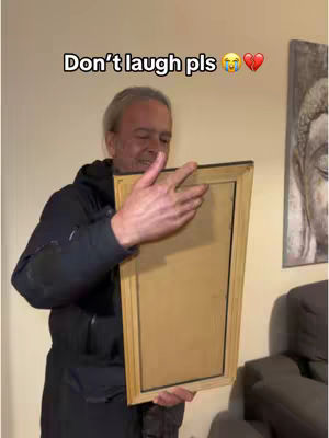 An older man with gray hair is holding a rectangular frame in front of him. He is smiling and looking slightly to the left. The background is a beige wall. A text overlay at the top says "Don't laugh pls" followed by a laughing emoji and a broken heart emoji. The man is wearing a dark jacket.