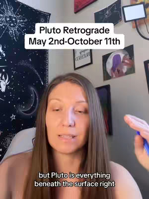 A woman with long brown hair is centered in the frame, looking directly at the camera. Behind her, a wall is decorated with various framed pictures and artwork. The overhead text reads 'Pluto Retrograde May 2nd-October 11th'. The overall tone is informative with the setting suggesting a personal space.