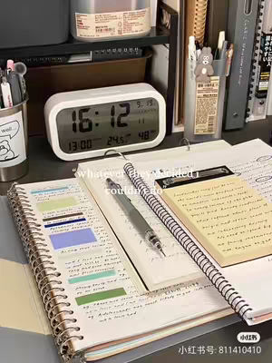 A close-up of a desk setup with a white digital clock displaying 16:12, a notebook with colorful sticky notes, a pen, and a pencil. The background includes a shelf with various items like pens and a container, creating a study or work atmosphere.