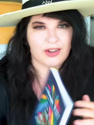 A woman with dark hair, wearing a hat and holding a tarot deck. She is looking at the camera, possibly talking about the deck.