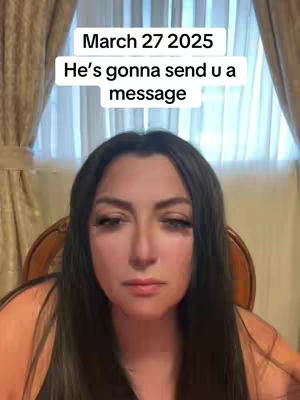 A woman with long dark hair stares into the camera with a neutral expression.  Text overlays read "March 27 2025" and "He's gonna send u a message". The background features curtains and a wooden chair.