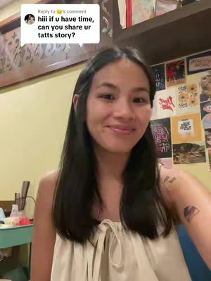 A woman with long black hair, smiling slightly, posed in front of a wall with various decorations on it. She wears a light-colored top and has a tattoo visible on her arm. A text box in the top-left corner has text reading, "hiii if u have time, can you share ur tatts story?"