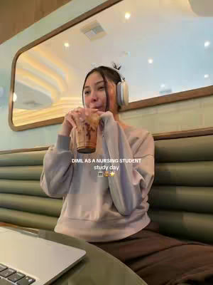 A young woman is seated in what appears to be a cafe or a similar setting, holding a beverage in her hands. She's wearing headphones and seems to be looking slightly off-camera with a contemplative or possibly slightly pensive expression. The background includes a section of green padded seating and a large mirror. The upper part of the image is brighter than the lower half. A laptop is partially visible, suggesting a study or work environment.
