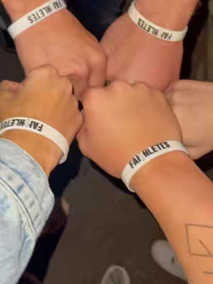 An overhead shot shows four hands clasped together, making a circle. Each wrist holds a white band with the words "FAITHLETES" printed on it. The hands appear to be of varying skin tones. The background and lighting are low-key, suggesting an indoor setting.