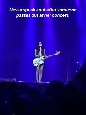 The image shows a woman on a stage holding a guitar, under blue spotlights. Text overlayed says 'Nessa speaks out after someone passes out at her concert!' with the woman in the center, stage and audience are visible.