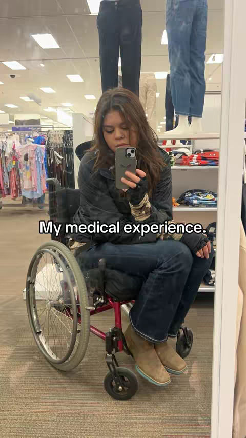 A woman in a wheelchair is taking a selfie in front of a mirror at what appears to be a store, likely a clothing retailer. She is using her phone to take the picture. The reflection shows the inside of the store. In front of the woman are racks with clothes, and mannequins standing with clothes behind her. The woman is wearing a dark colored jacket, dark pants, and boots.