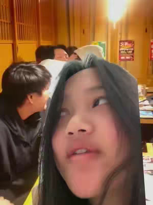 A selfie-style shot focuses on a young woman with long black hair, gazing slightly upwards and to her right. She's in the foreground, while out-of-focus figures and a warm-toned, likely indoor setting are visible in the background. The lighting appears to be natural or from overhead sources.