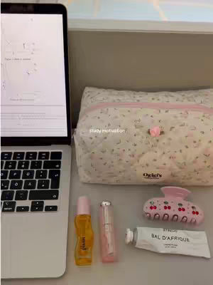 The image showcases a neatly organized study setup on a light-colored surface. A laptop displaying a document, a floral-patterned makeup bag, a pink hair clip shaped like cherries, two perfume bottles, and a tube of lip balm are arranged. The color scheme is soft and feminine, with a focus on pinks and whites. The laptop is positioned on the left, appearing open and in use.