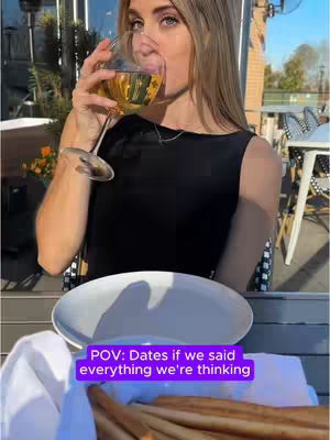 A woman is shown seated at a table, drinking from a wine glass, with a plate and breadsticks in the foreground. The background is a sunny outdoor setting with trees and other tables.