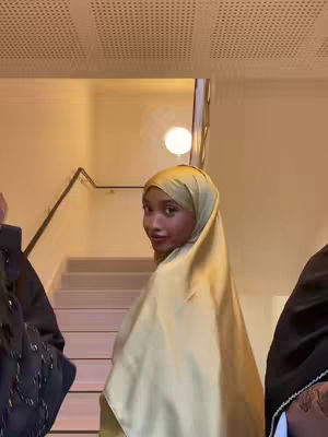 A woman wearing a gold hijab turns to look over her shoulder at the camera. She is standing inside a building that has cream-colored walls, a staircase, and a decorative ceiling.