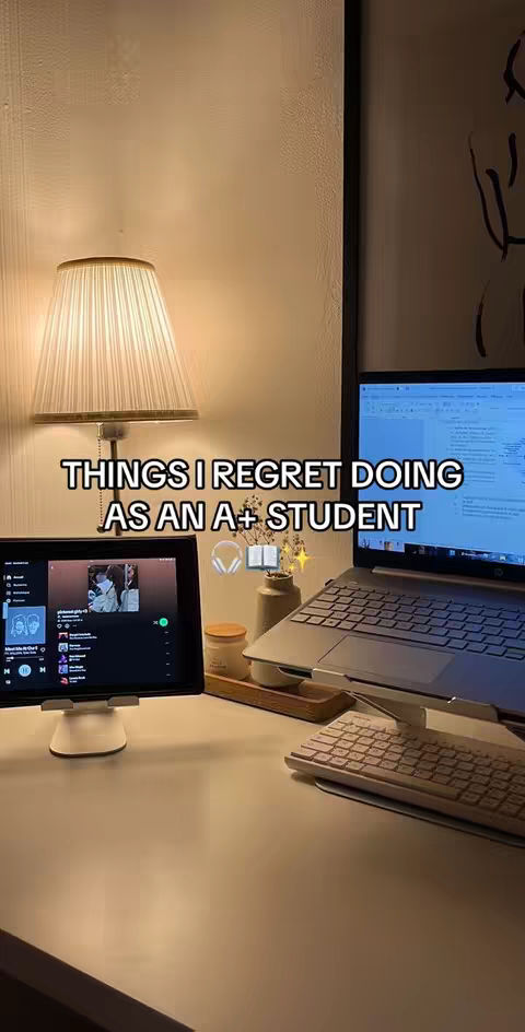 A well-lit workspace featuring a laptop, a tablet displaying a music app, desk accessories, and a lamp. Text overlayed on the image reads "THINGS I REGRET DOING AS AN A+ STUDENT."