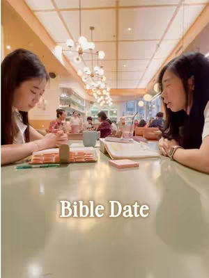 The image captures a moment of shared intimacy and spiritual practice within a cozy, well-lit cafe setting. Two young women, positioned on either side, are seen leaning over a table. Each woman is intently focused on an open book, with one, further away, having their hands clasped in a position of prayer or reflection. The table is a light green color. In the background, other patrons are subtly blurred.