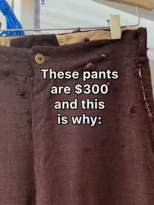The image shows a close-up of a pair of worn, dark brown pants hanging on a clothes hanger. The pants have visible holes and signs of wear and tear throughout the fabric. The top of the pants, including the waistband and a button, are visible. Text overlayed on the image reads, "These pants are $300 and this is why:"
