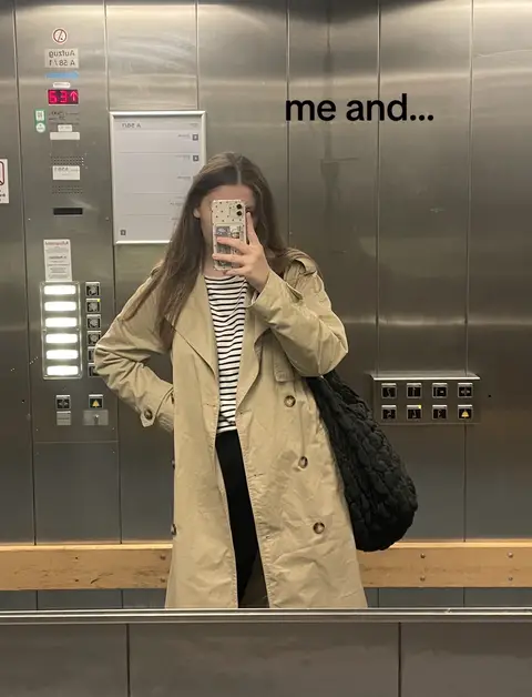 A woman in a beige trench coat stands in a mirrored elevator, holding a phone in front of her face, partially obscuring her features. The lighting is bright, the overall composition simple, with muted colors from the coat and elevator interior.