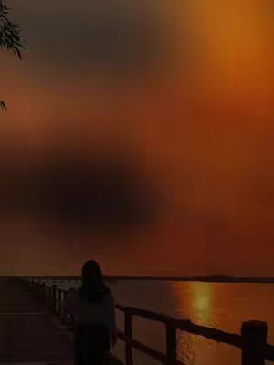 The image captures a serene sunset scene. Silhouetted against a vibrant sky, a person walks away from the camera along a wooden walkway that extends over water reflecting warm orange and yellow hues. The sky fills most of the frame, creating a sense of peace and reflection.