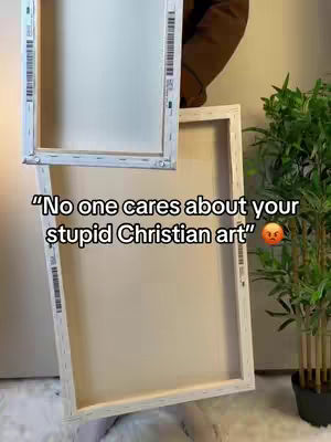 The image shows two canvas frames. The bottom frame is angled toward the viewer, and the top frame is held slightly above. Text overlay reads "No one cares about your stupid Christian art" with an angry emoji. In the background are someone's arms and a green plant in a pot.