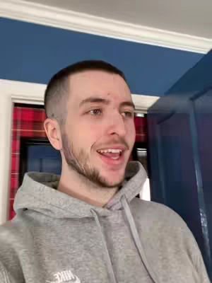 A man in a gray hoodie is smiling and looking slightly off-camera. The background includes a blue wall and a partially visible door frame with a plaid pattern.