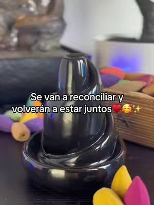 The image presents a close-up of an aromatherapy fountain with a black, glossy finish set against a blurred background. The fountain has a central, spiraling design, with a lower circular basin and a series of colorful orbs. Text in white overlay reads, 'Se van a reconciliar y volverán a estar juntos', with emoji of hearts, a smiling face and stars.